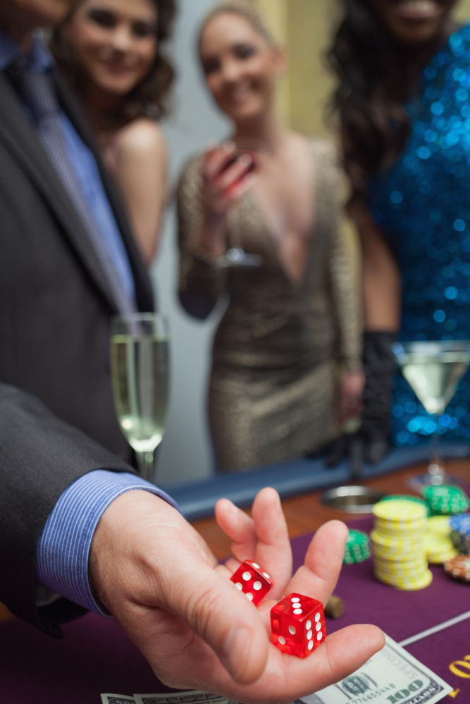 Man holding dice at a playing table with people, drinks and casino chips in the background